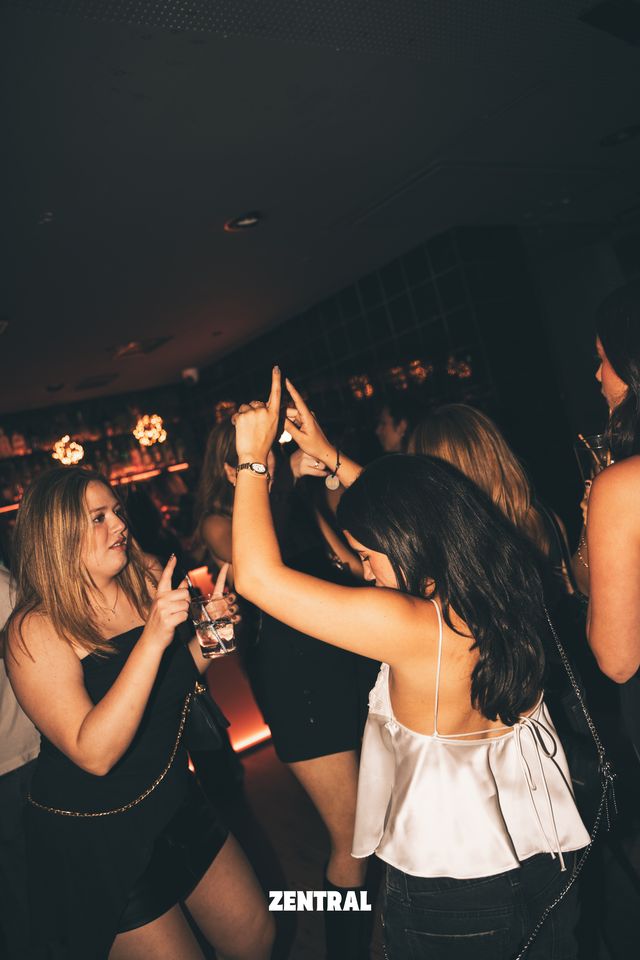 Dos chicas bailan junto a la barra, una de negro con una copa y otra de espaldas con top de satén blanco y las manos en alto, bajo luces cálidas.
