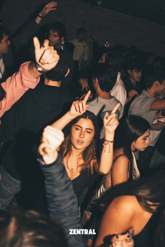 Una chica de pelo largo baila con los dos brazos en alto y los dedos señalando arriba, rodeada de chicos y chicas en la pista.
