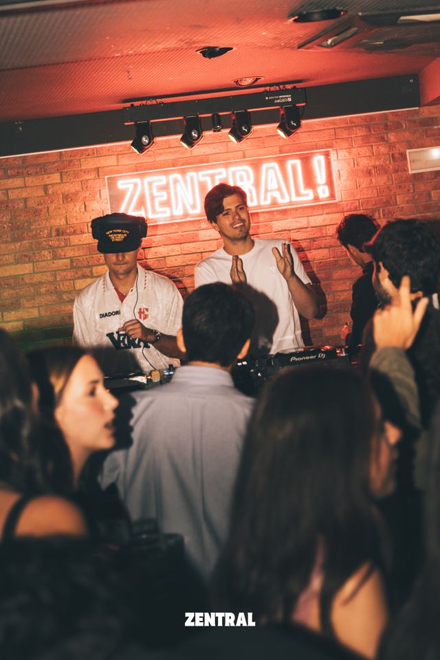 Un DJ con gorra mezcla mientras un chico sonriente aplaude a su lado, bajo el neón Zentral y varios focos, con el público en primer plano.