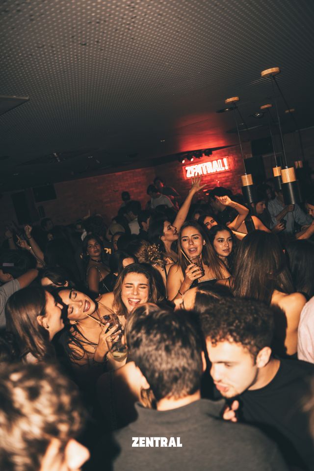 Chicas y chicos bailan apretados en la pista entre risas, con copas en la mano, bajo el neón rojo de Zentral y la pared de ladrillo.