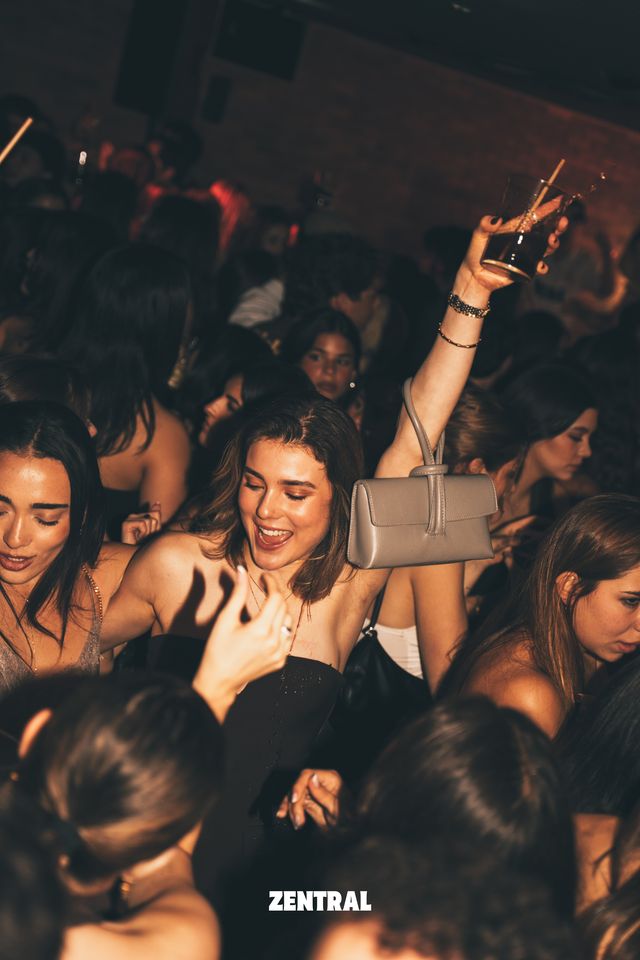 Una chica sonríe en medio de la multitud mientras un brazo en alto sostiene una copa con pajita y lleva un bolso gris colgado.