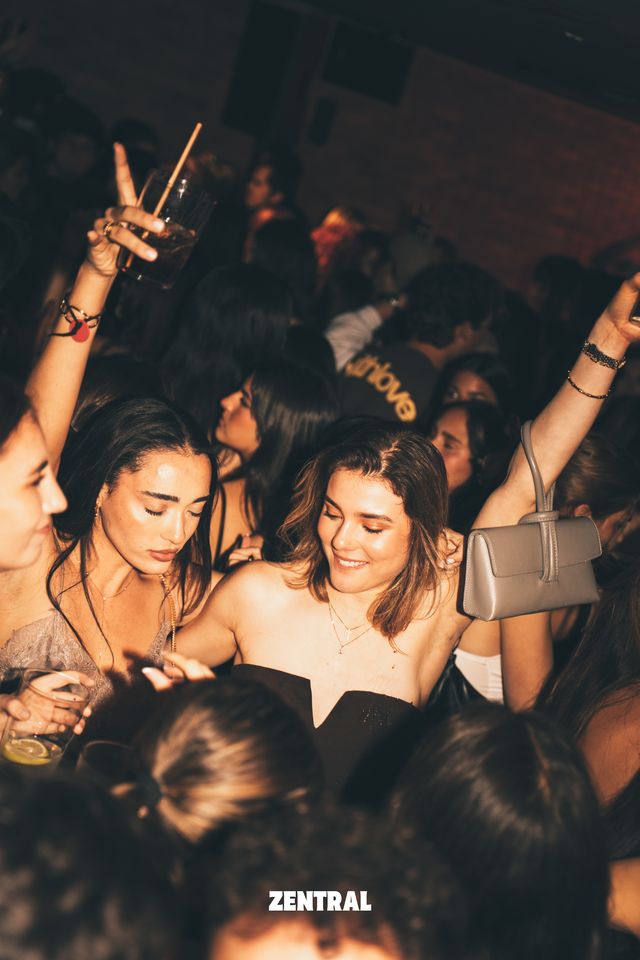 Dos chicas miran hacia abajo entre la multitud, una de ellas sonriendo, rodeadas de brazos en alto con una copa y un bolso gris colgado.
