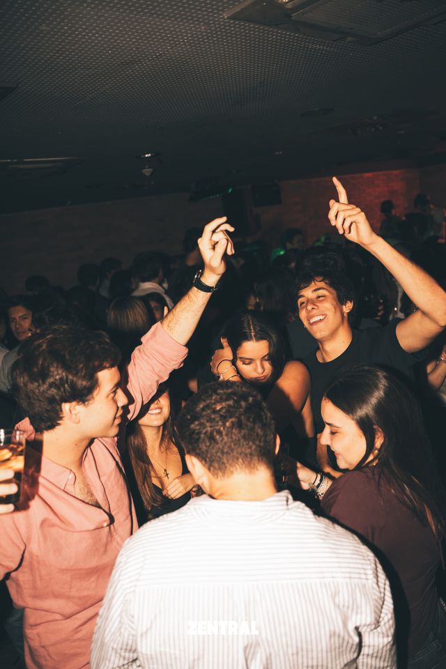 Un grupo de chicos y chicas ríe y baila con los brazos en alto en una sala oscura, con un chico de camisa rosa a la izquierda.