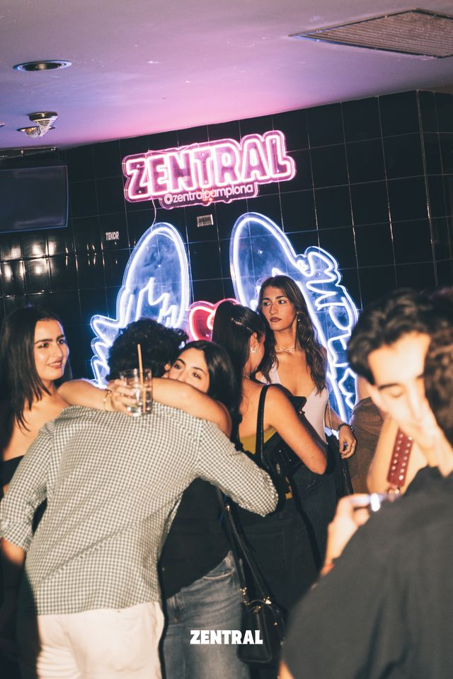 Un chico de camisa de cuadros abraza a una chica en primer plano, junto al neón rosa de Zentral y unas alas de ángel luminosas.