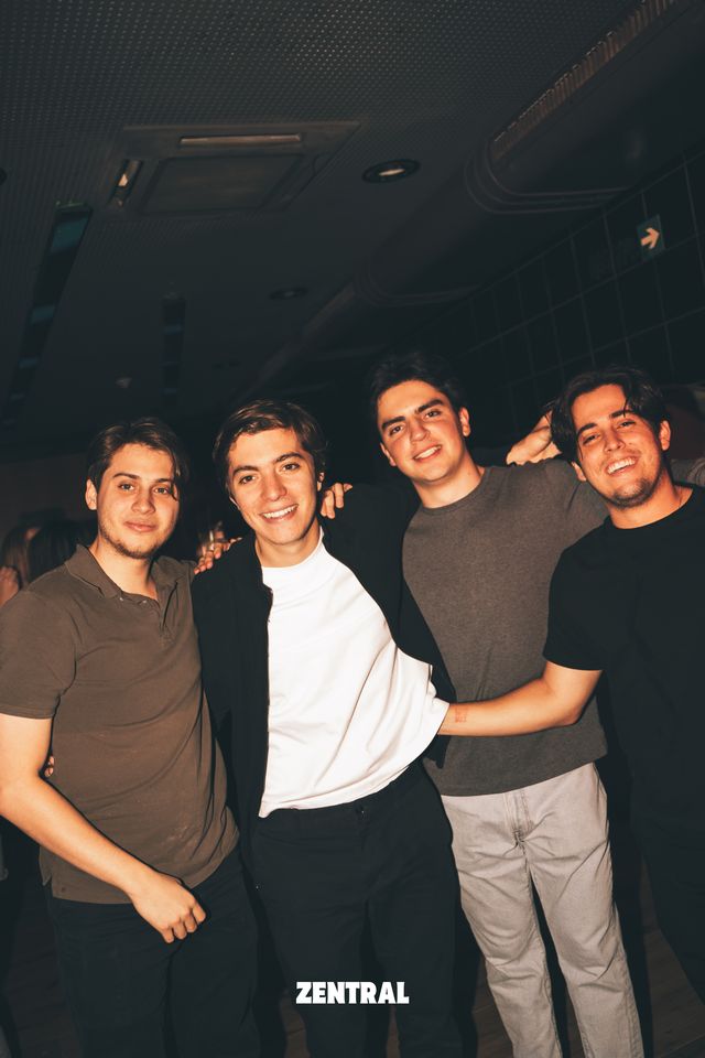 Cuatro chicos posan abrazados y sonriendo a cámara en la sala del club, con el techo oscuro y una pared de azulejos detrás.