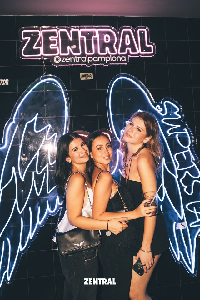 Tres amigas posan abrazadas ante el neón rosa de Zentral y unas grandes alas de ángel luminosas, una de ellas con una copa.