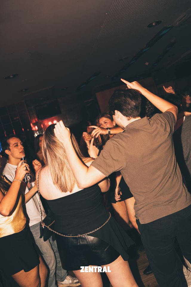 Una chica rubia, de espaldas, y un chico de polo marrón con un brazo en alto bailan entre la gente en una sala en penumbra.