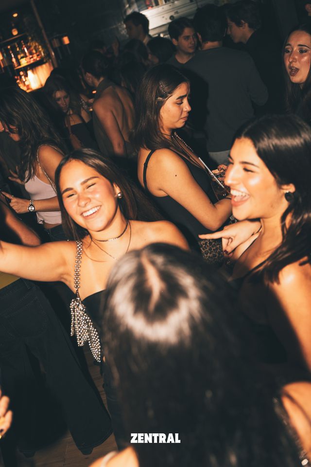 Dos chicas bailan y se ríen a carcajadas en plena pista, con más gente alrededor y una barra iluminada al fondo.