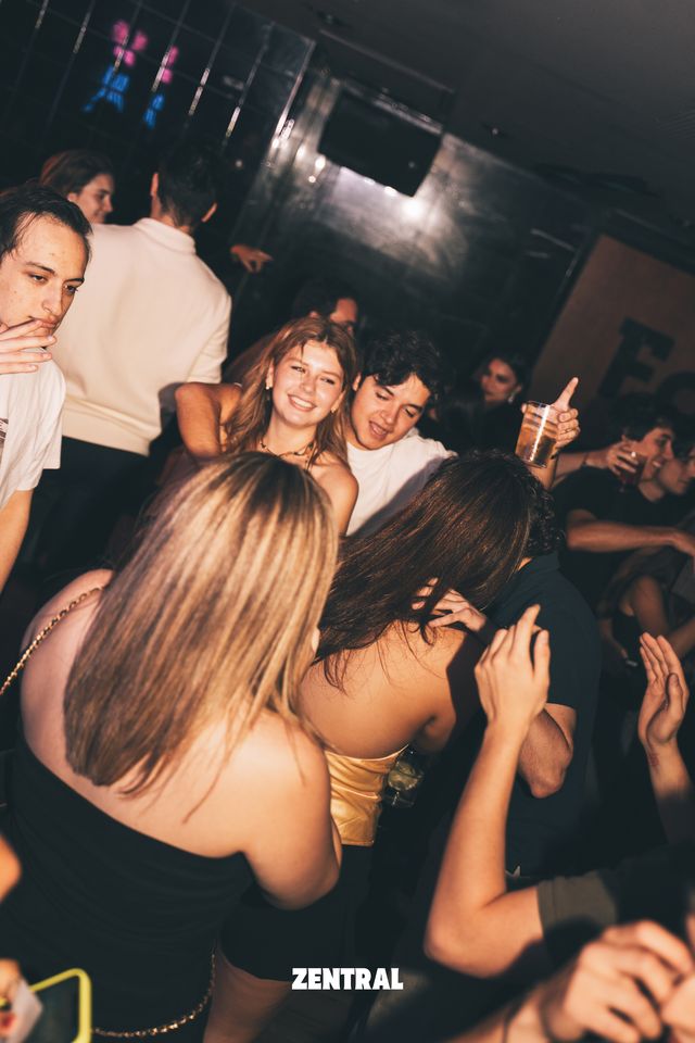 Una chica y un chico sonríen entre la multitud, con dos chicas bailando de espaldas en primer plano y una pared oscura detrás.
