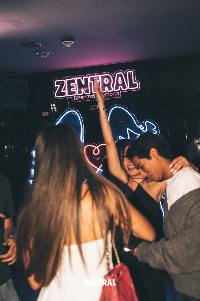 Una chica de espaldas con bolso rojo en primer plano; detrás, otra baila con el brazo en alto abrazada a un chico, bajo el neón de Zentral.