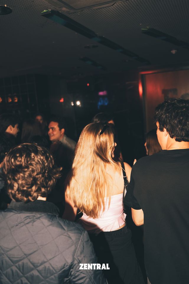 Una chica de pelo largo y rubio, de espaldas, entre un chico de pelo rizado y otro con camiseta negra, en una sala oscura llena de gente.