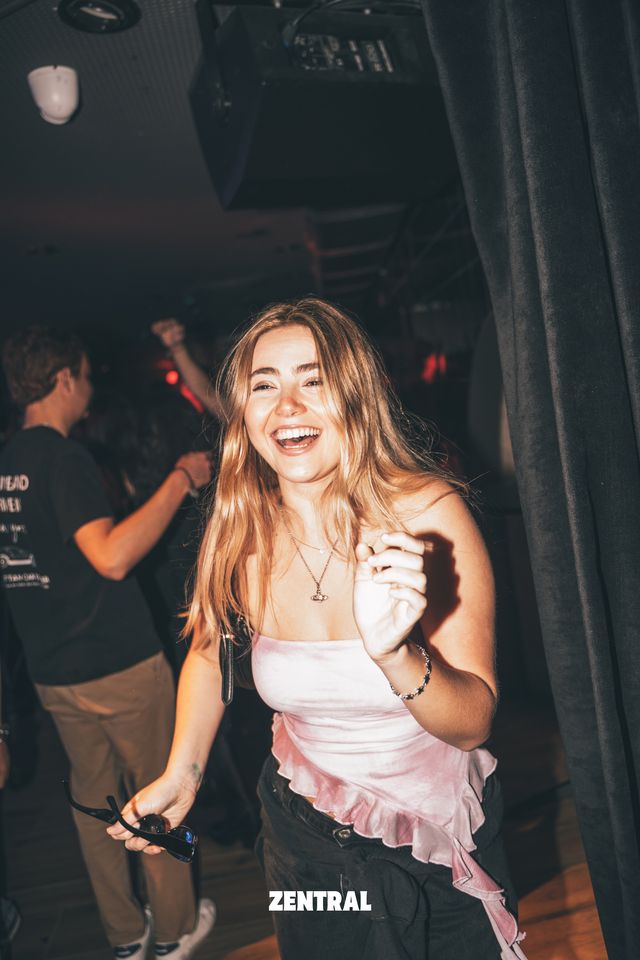Una chica ríe con ganas mientras baila con unas gafas de sol en la mano, junto a una cortina oscura y un chico al fondo.