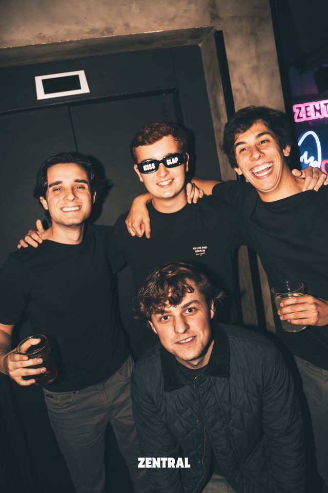Cuatro chicos posan juntos sonriendo a cámara, uno con gafas de sol negras y otros dos con un vaso en la mano, ante una pared oscura.