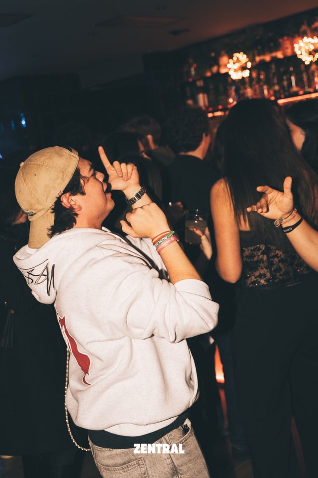Un chico con gorra beige y sudadera blanca baila señalando hacia arriba, con la barra de un bar y luces cálidas al fondo.
