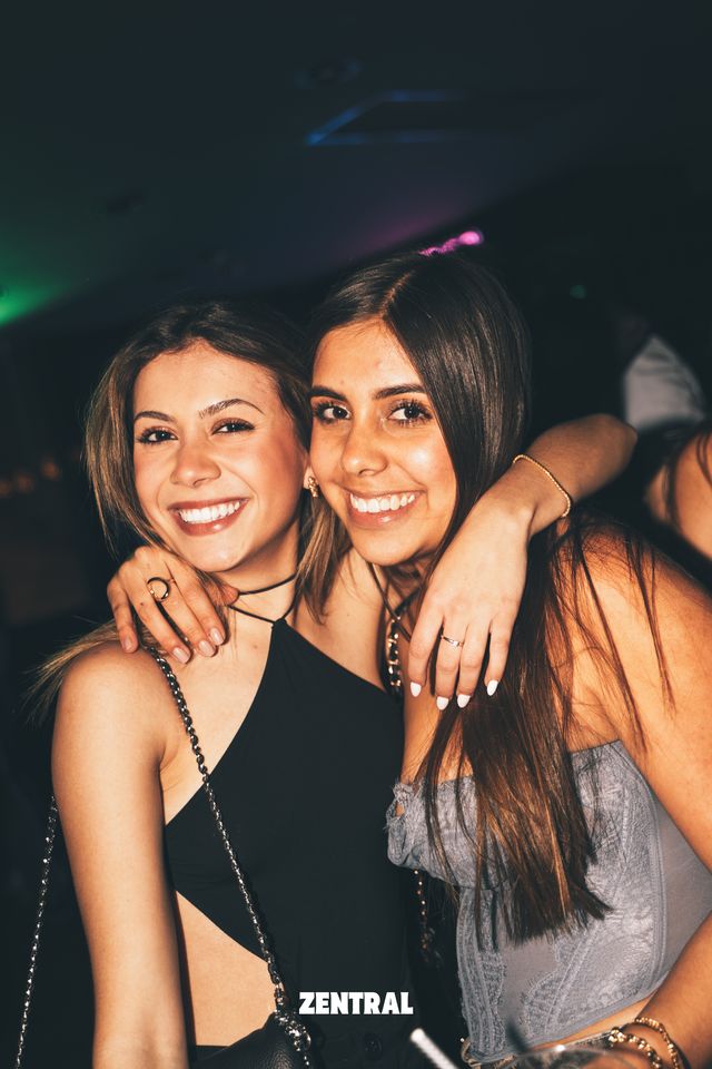 Dos chicas posan abrazadas sonriendo a cámara, una con top negro y otra con corsé gris, en una sala oscura con luces verdes y rosas.