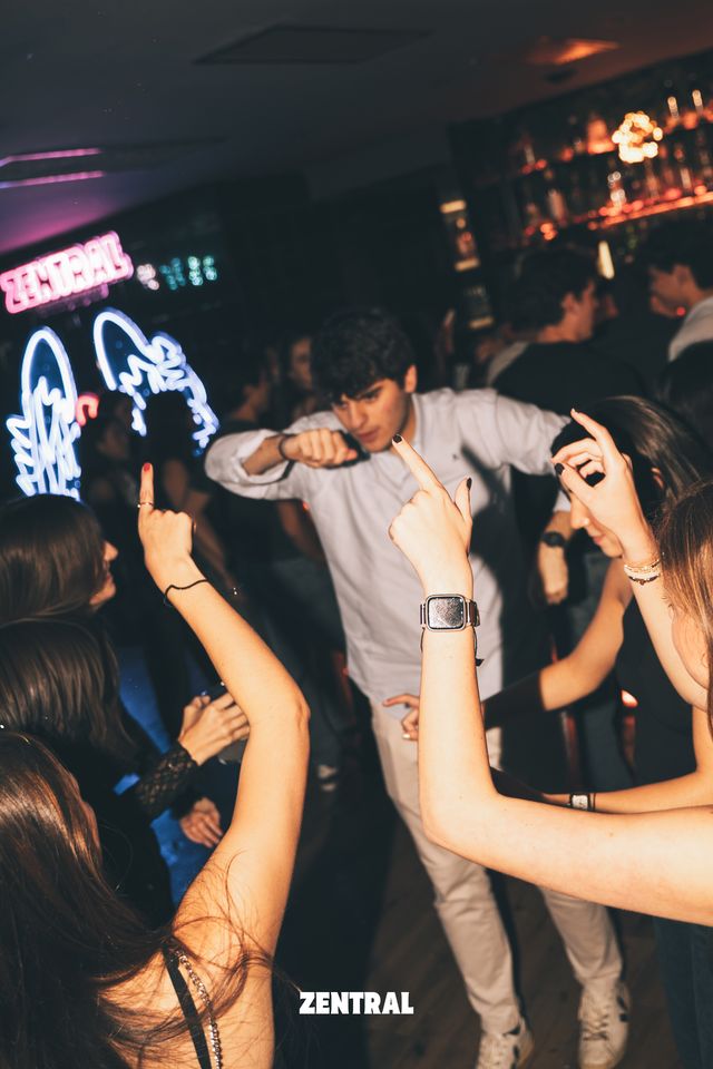 Un chico con camisa clara baila entre chicas que levantan las manos, con neones azules y rosas a la izquierda y la barra del bar al fondo.