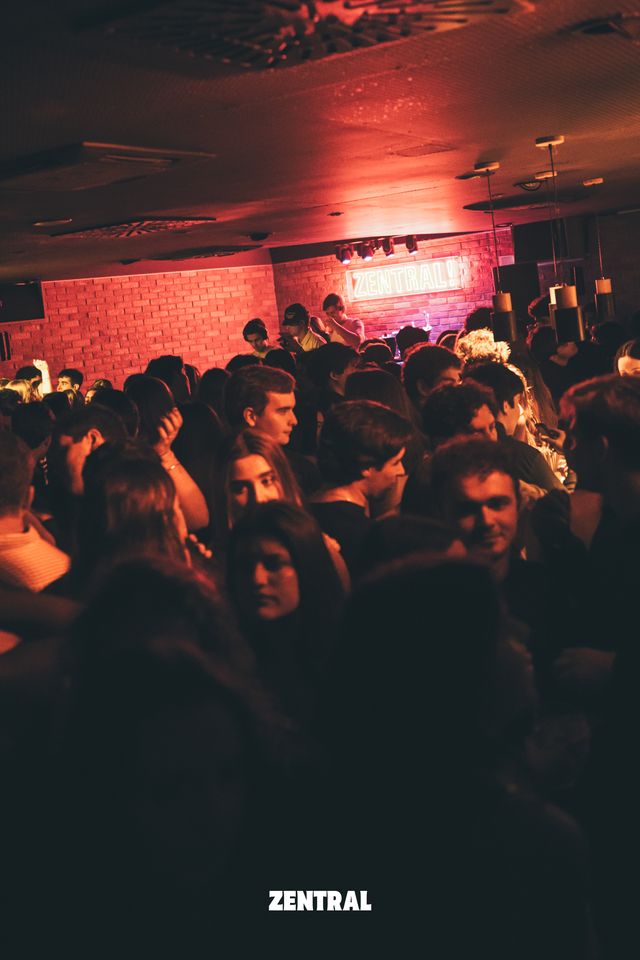 Una sala abarrotada de gente bajo una luz roja, con la cabina del DJ, la pared de ladrillo y el neón de Zentral al fondo.