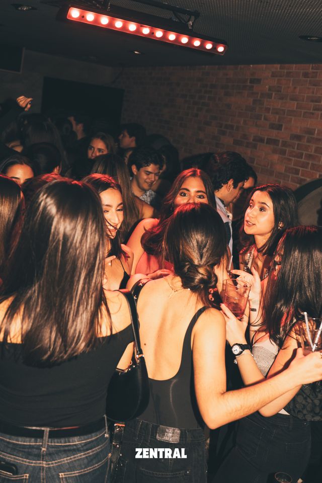 Un grupo de chicas charla con vasos en la mano entre la multitud, bajo una barra de luces rojas y con una pared de ladrillo detrás.