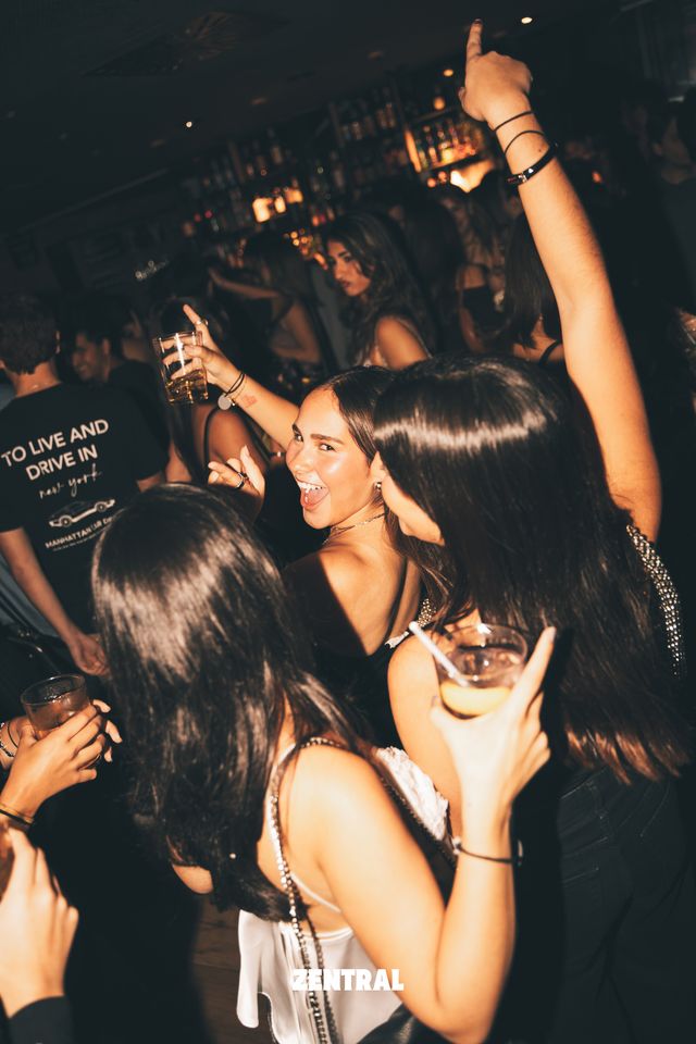 Un grupo de chicas baila con copas en la mano y un brazo en alto; una mira a cámara sonriendo, con las botellas de la barra al fondo.