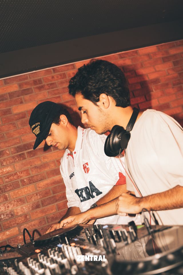 Dos DJs mezclan juntos frente a una pared de ladrillo, uno con gorra negra y camiseta blanca y otro con auriculares al cuello.