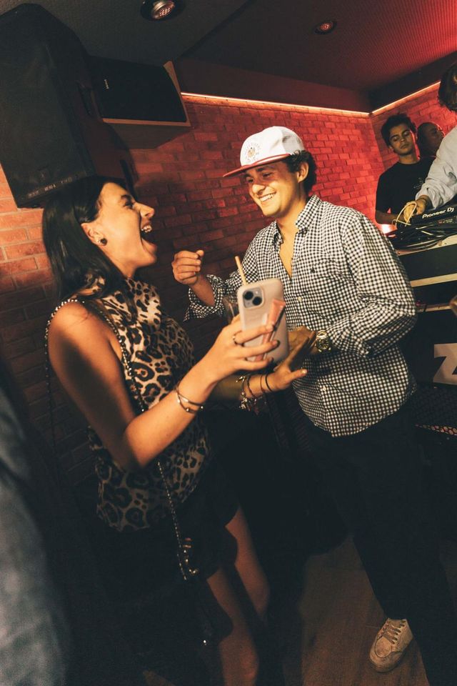 Una chica con top de leopardo ríe a carcajadas junto a un chico con gorra blanca y camisa de cuadros, al lado de la cabina del DJ.
