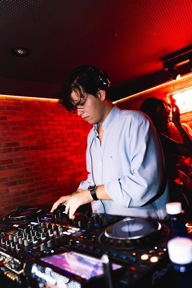 Un DJ con camisa celeste y auriculares mezcla en los platos bajo una luz roja, con la pared de ladrillo detrás.