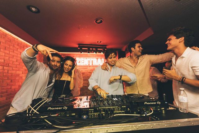 Un grupo de amigos celebra abrazado tras la mesa de DJ, bajo el techo rojo y el neón de Zentral.