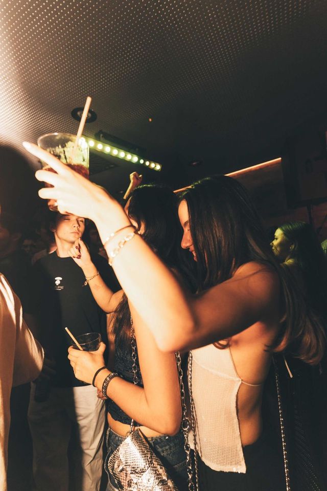 Dos chicas bailan muy juntas en la pista; una levanta una copa con pajita bajo luces verdes.
