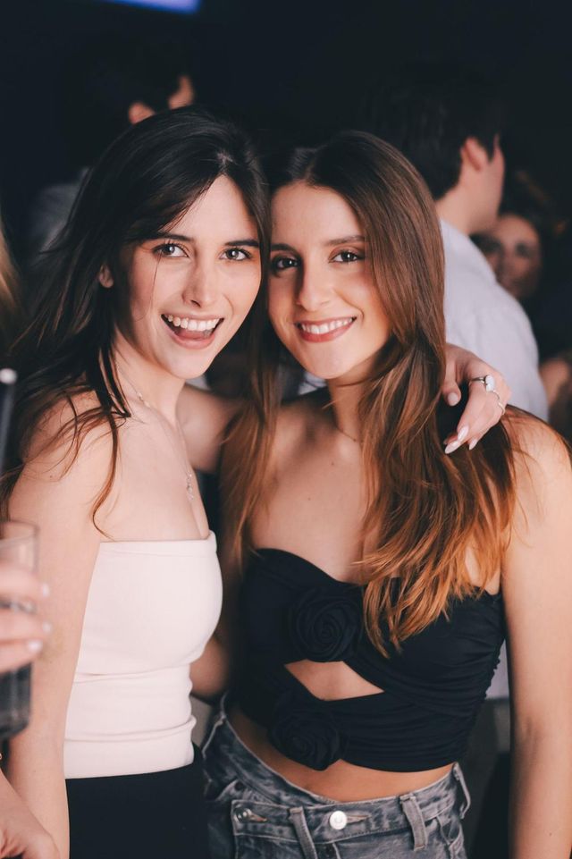 Dos amigas sonríen a cámara abrazadas; una con top blanco y otra con top negro con dos flores.