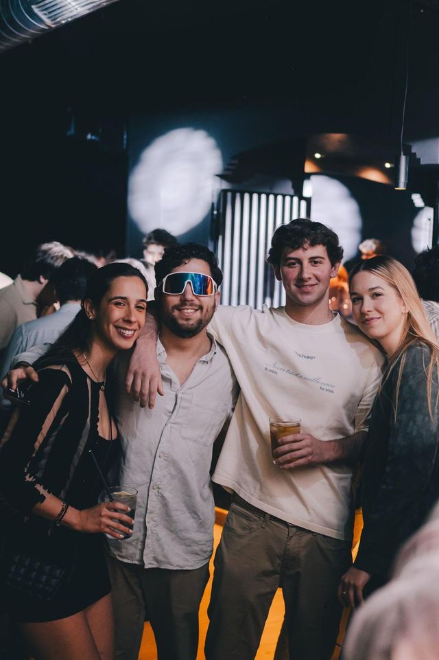 Cuatro amigos posan juntos y sonríen, dos de ellos con un vaso en la mano; otro lleva gafas de sol deportivas.