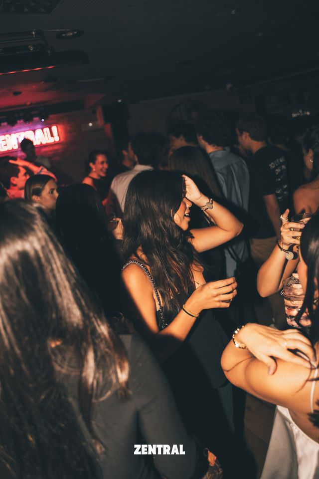 Una chica baila con la mano en el pelo entre la multitud, en una sala en penumbra con el neón de Zentral al fondo a la izquierda.