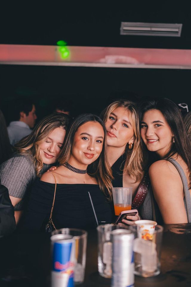 Cuatro amigas posan muy juntas en la barra, con vasos y latas delante.