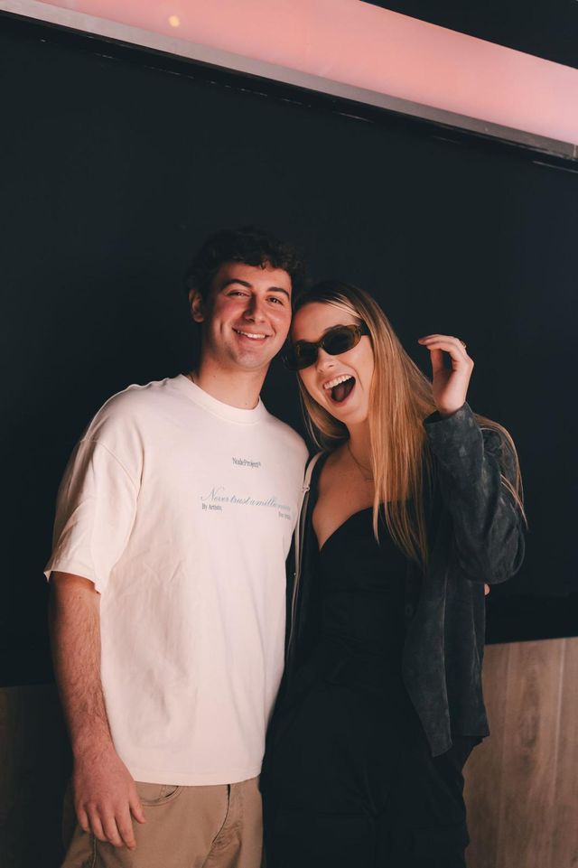 Una pareja posa sonriente ante un fondo negro: él con camiseta blanca y ella con gafas de sol, riendo.