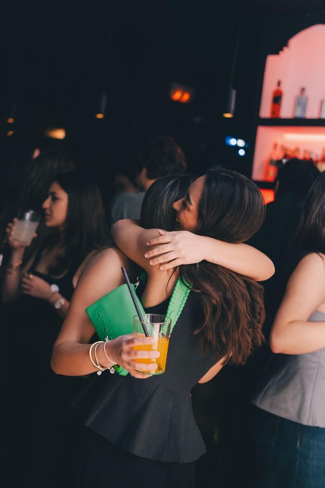 Dos amigas se abrazan entre la gente; una sostiene una copa naranja y un bolso verde, con la barra al fondo.