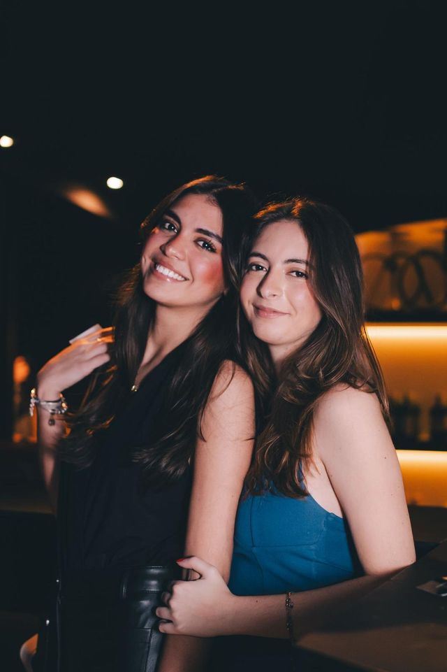 Dos amigas posan sonrientes junto a la barra iluminada en naranja; una con top azul.