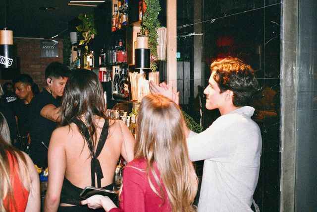 Gente pidiendo en la barra, de espaldas, mientras un camarero sirve tras el mostrador lleno de botellas.