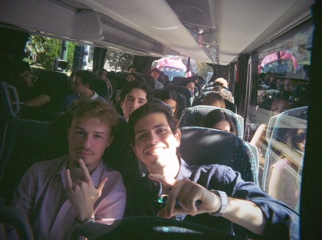 Dos chicos hacen gestos con las manos en el autobús, uno con una gran sonrisa; detrás, los asientos van llenos.
