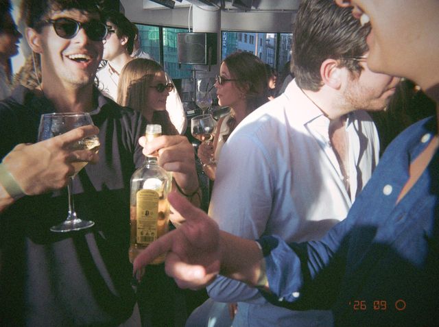 Un chico con gafas de sol se ríe con una copa de vino mientras alguien sostiene una botella en la sala soleada.