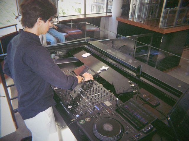 Un DJ con camisa azul marino y gafas pincha en la mesa de mezclas mientras entra la luz del día por las ventanas.