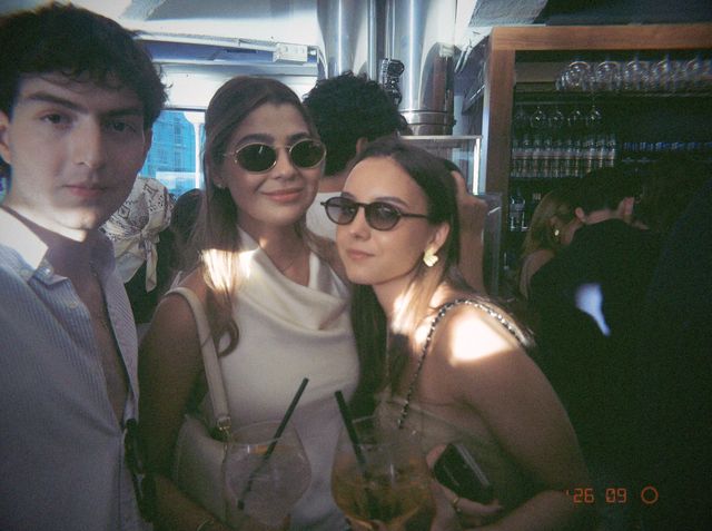 Dos chicas con gafas de sol y copa en mano posan junto a un chico en la barra, con manchas de sol en los hombros.