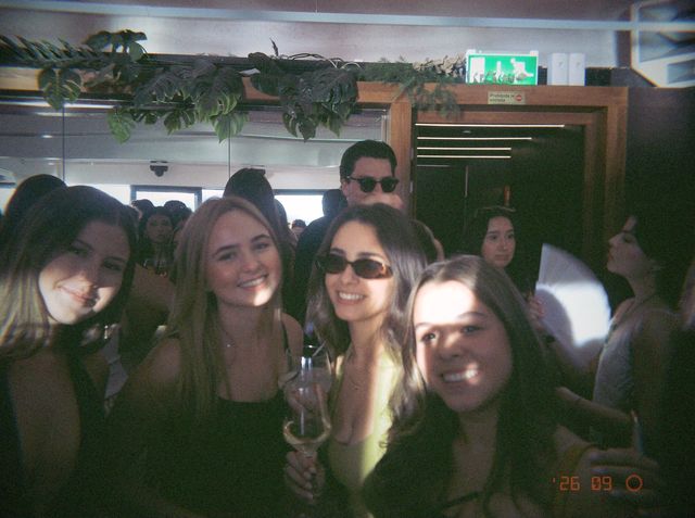 Cuatro amigas sonríen apiñadas, una con gafas de sol y una copa, bajo una guirnalda de hojas junto a una puerta.