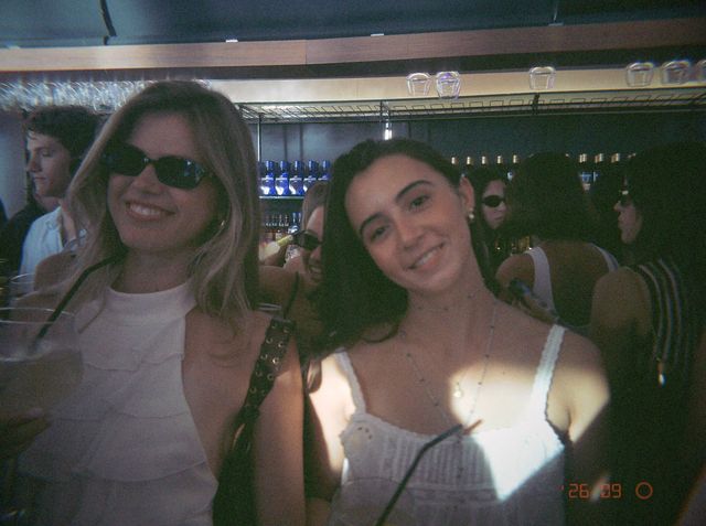 Dos chicas vestidas de blanco sonríen en la barra, una con gafas de sol y otra con una copa con pajita.