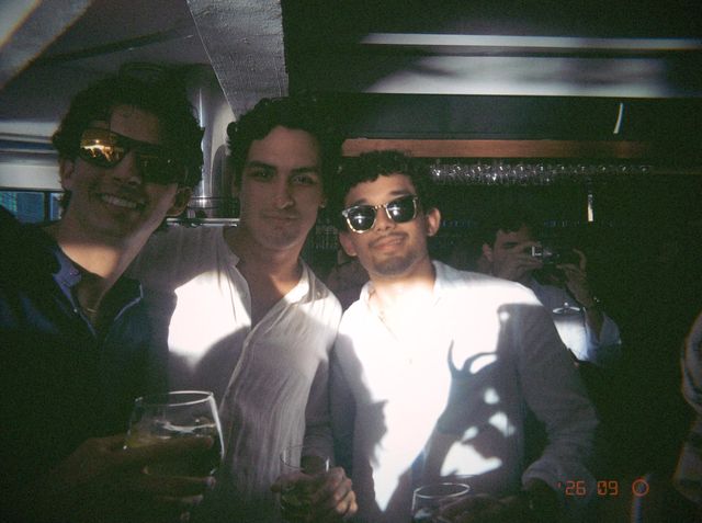 Tres chicos, dos con gafas de sol, posan copa en mano junto a la barra con la sombra de unas hojas en la camisa.