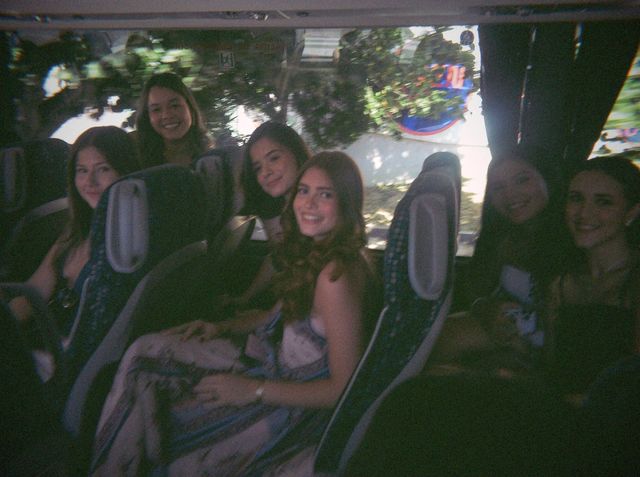 Seis chicas sonríen desde los asientos del autobús; delante, una con un vestido estampado en azul y blanco.