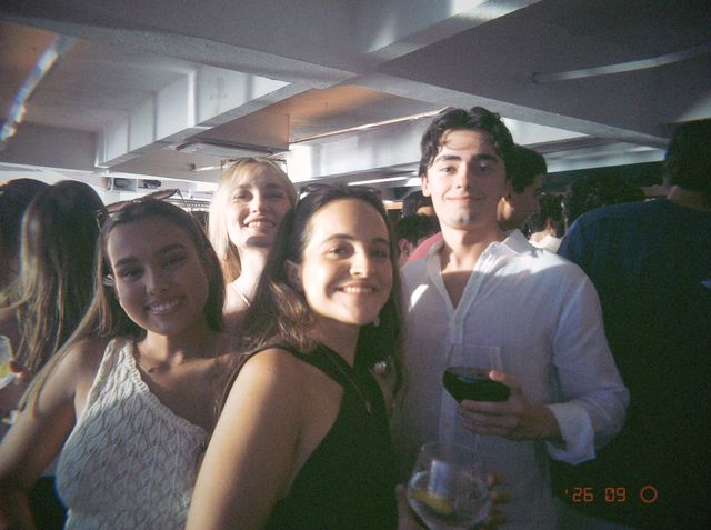 Cuatro amigos sonríen juntos; un chico con camisa blanca sostiene una copa de vino y delante asoma otra con hielo.