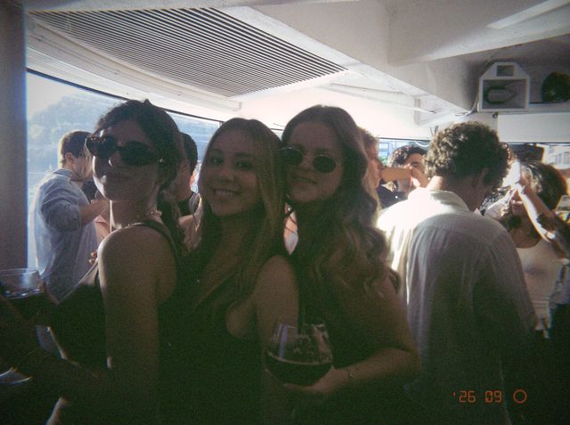 Tres amigas posan juntas con una copa de vino, dos de ellas con gafas de sol, delante de los ventanales luminosos.
