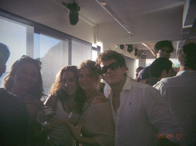 Tres chicas, dos con vaso en la mano, y un chico con gafas de sol posan muy juntos al lado de un ventanal soleado.