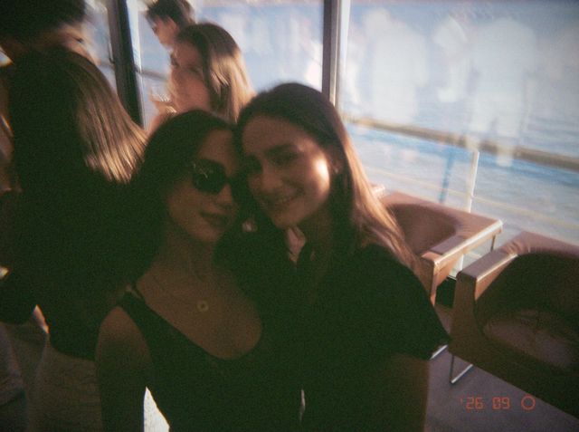 Dos amigas juntan las mejillas, una con gafas de sol oscuras, junto a los ventanales que dan al mar.