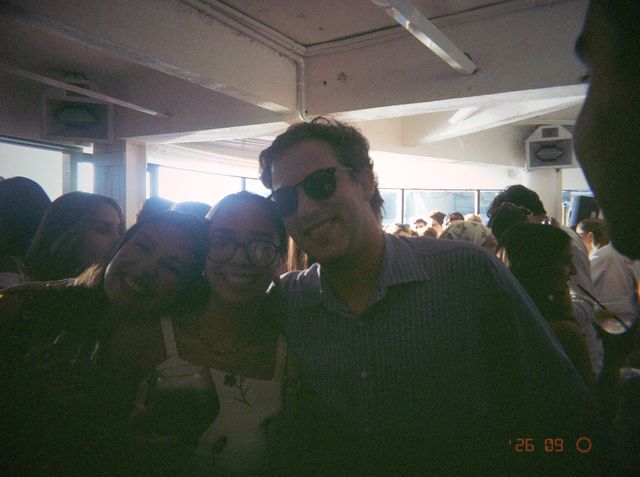 Dos chicas, una con gafas, se arriman sonrientes a un chico con gafas de sol y camisa de rayas en el local lleno.