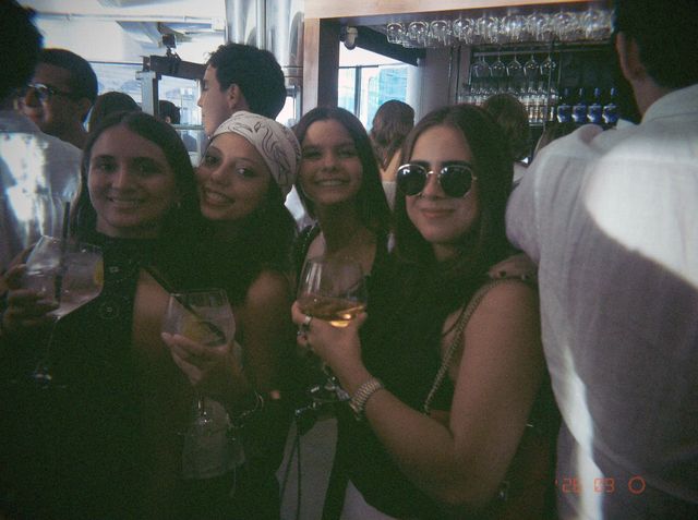 Cuatro amigas posan con copas en la barra; una lleva un pañuelo en la cabeza y otra, gafas de sol redondas.